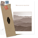 The Mountains Thank You Card by Shannon Martin Design features "Mountains of gratitude" in gold over sepia mountain ranges, paired with a brown envelope and vintage stamps. Made in the USA from recycled paper.