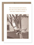 A sepia-toned image reminiscent of vintage photos features a woman in a white swimsuit, smiling as she hangs from a tree with both hands and one leg. Above, the text reads: "You never know how strong you are until being strong is the only choice you have." This artwork is part of the Strong Encouragement Card collection by Shannon Martin Design.