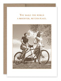 Sepia-toned image of two women smiling with a tandem bicycle in a grassy field. The text reads, "You make the world a brighter, better place." This card from the Shannon Martin Design collection is elegantly paired with a brown recycled paper envelope. Product: A Better Place Thank You Card.