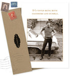 The Handsome Birthday Card by Shannon Martin Design features a photo of a man in stripes and jeans by a car with the message, "It’s tough being both handsome and humble." Includes an envelope with vintage-style stamps. Made in USA.