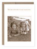 The Shenanigans Birthday Card by Shannon Martin Design features a sepia-toned photo of two children smiling while lying on grass. The card displays the text, "You're the she to my nanigans," above them. It's crafted with recycled paper in the USA and comes with a simple white border paired with a brown envelope.