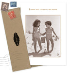 The I Wish You Lived Next Door Friendship Card by Shannon Martin Design features two girls in swimsuits holding hands on a beach, with a brown envelope and old stamps—perfect as a blank greeting card for any occasion.