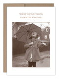 The Under The Weather Get Well Card by Shannon Martin Design features a vintage-style, recycled paper design of a girl in a coat holding an umbrella and looking up at the sky with an open hand. The text reads, Sorry you‚Äôre feeling under the weather, set against a simple outdoor background.