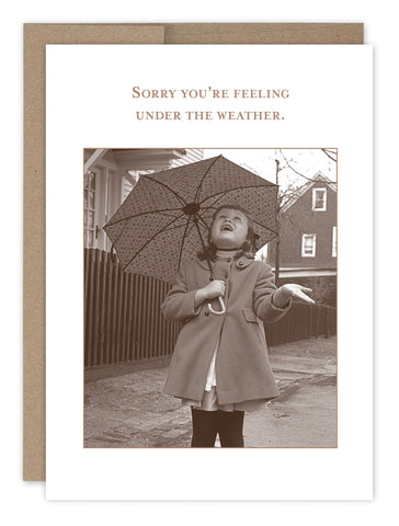 The Under The Weather Get Well Card by Shannon Martin Design features a vintage-style, recycled paper design of a girl in a coat holding an umbrella and looking up at the sky with an open hand. The text reads, Sorry you’re feeling under the weather, set against a simple outdoor background.