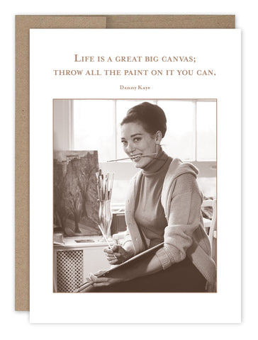 A woman smiles with paintbrushes, one in her mouth, in front of a painting on recycled paper. The Big Canvas Birthday Card by Shannon Martin Design features a vintage photo and a Danny Kaye quote: Life is a great big canvas; throw all the paint on it you can. Made in the USA creatively.