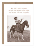 A sepia-toned image from Shannon Martin Designs collection shows a person wearing a cowboy hat, playing guitar while standing on a horse in a grassy field. The Dont Stop Laughing Birthday Card reads: You dont stop laughing because you grow old; you grow old because you stop laughing.