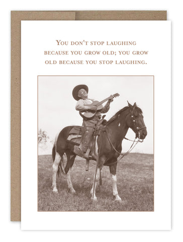 A sepia-toned image from Shannon Martin Designs collection shows a person wearing a cowboy hat, playing guitar while standing on a horse in a grassy field. The Dont Stop Laughing Birthday Card reads: You dont stop laughing because you grow old; you grow old because you stop laughing.