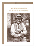 A smiling person in a hat and vest holds two puppies with the text “The best things in life are meant to be held close” above. This sepia-toned image appears on Shannon Martin Design’s Best Things In Life Birthday Card, made in the USA.