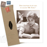 The My Turn Birthday Card by Shannon Martin Design features a vintage-style woman reading on her stomach with the message, "This chapter of my life is called My Turn." Includes envelope with old postage stamps. Made in the USA.