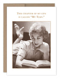 The My Turn Birthday Card by Shannon Martin Design features a black-and-white photo of a woman reading, with the text “This chapter of my life is called My Turn.” Elegant design with a white border. Made in the USA.