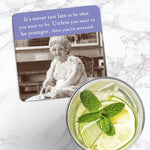 The Never too late Coaster by Shannon Martin Design, featuring an elderly woman and witty text, rests beside lemon-mint water on a marble surface.