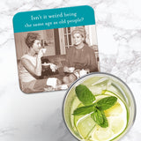 The Shannon Martin Design "Same Age Coaster," featuring a vintage photo of two women and the text "Isn't it weird being the same age as old people?" is shown cork-backed on marble, next to a glass of lemon water with mint.