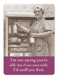 An older woman in glasses and an apron smiles, holding a milk bottle by an open fridge. Caption: "I’m not saying you’re old, but if you were milk, I’d sniff you first." Cream Of The Crop Magnet by Shannon Martin Design—perfect for vintage photo fans.