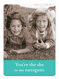 Two girls smile on the grass; one with curly hair sticks out her tongue. Text below reads, "You're the she to my nanigans." The Shannon Martin Design Shenanigans Magnet brings playful charm to your kitchen décor.
