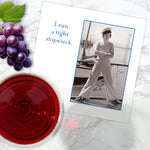 A Tight Shipwreck Napkin by Shannon Martin Design sits on a marble surface alongside red wine, grapes, and a greeting card featuring a person in nautical attire with the phrase "I run a tight shipwreck.