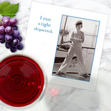 A Tight Shipwreck Napkin by Shannon Martin Design sits on a marble surface alongside red wine, grapes, and a greeting card featuring a person in nautical attire with the phrase "I run a tight shipwreck.