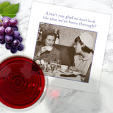 The Shannon Martin Design "Aren't you Glad Napkin" features a black-and-white photo of two women talking at a restaurant, alongside grapes, red wine, and napkins. The card reads: Aren’t you glad we don’t look like what we’ve been through?.