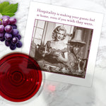 A glass of red wine and grapes rest beside a card on Shannon Martin Design’s Hospitality Napkin, featuring a vintage photo of a woman serving tea. The card reads: Hospitality is making your guests feel at home, even if you wish they were.