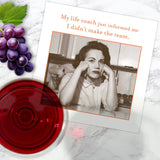 A Shannon Martin Design "Life Coach Napkin" lies on a marble surface next to red wine and grapes. The napkin features a vintage photo of a disappointed woman and the text: "My life coach just informed me I didn’t make the team.