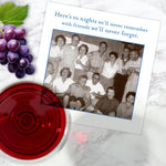 A Shannon Martin Design "Here's To Nights" napkin lies on a marble surface beside grapes, red wine, and a vintage-style greeting card featuring laughing friends and the message: "Here's to nights we'll never remember with friends we'll never forget.