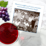 A Shannon Martin Design "Here's To Nights" napkin lies on a marble surface beside grapes, red wine, and a vintage-style greeting card featuring laughing friends and the message: "Here's to nights we'll never remember with friends we'll never forget.