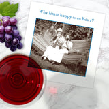The Happy Hour Napkin by Shannon Martin Design is shown on marble beside grapes, a glass of red wine, and a card featuring two women on a hammock with drinks and the phrase, "Why limit happy to an hour?".