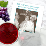 A Shannon Martin Design “Kind Of Crazy Napkin” rests on marble next to red wine, grapes, and a photo card reading, "Life is all about finding people who are your kind of crazy," with three people in costume.