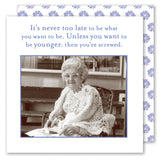 An elderly woman in glasses and a crocheted shawl sits at a table with Shannon Martin Design’s Never Too Late Napkin. Above her: It’s never too late to be what you want. Unless you want to be younger—then you’re screwed.