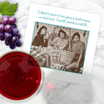 The Shannon Martin Design "Glass Half Full Napkin" is shown on a marble surface with a greeting card featuring three women at a bar, a glass of red wine, and grapes. Card reads: I don’t care if the glass is half empty or half full. I still need a refill.