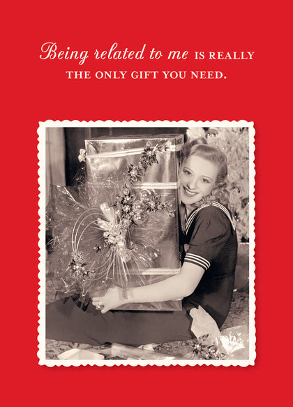 A vintage-style black-and-white photo showcases a smiling person holding a large gift wrapped in recycled paper. The background features a red hue with decorative white text, "Being related to me is really the only gift you need," from the *Being Related Holiday Card* by Shannon Martin Design.