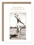 A cowboy showcases an impressive stunt, balancing on one leg atop a white horse while waving his hat. Above him, the text reads: Live your life and forget your age. Norman Vincent Peale. The image is framed with a brown vintage photo style and features a blank card made of recycled paper in the background, part of Shannon Martin Design's "Live Your Life Birthday Card" collection.