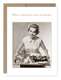 The What A Friend Birthday Card by Shannon Martin Design features a vintage-style illustration of a smiling woman with short, curled hair, wearing a sleeveless, collared dress. She is holding a tray filled with various cheeses. The card is printed on recycled paper and includes the text "What a friend we have in cheeses" at the top. Made in the USA.