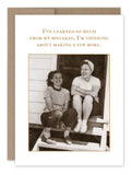 Two women sit on porch steps, smiling. One is dressed in a striped shirt and pants, while the other wears a headscarf, t-shirt, shorts, and socks. A Shannon Martin Design card titled "My Mistakes Birthday Card" is displayed above them with the message: ‚ÄúI've learned so much from my mistakes, I'm thinking about making a few more,‚Äù printed on recycled paper for an eco-friendly touch.