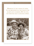 A vintage-style photo captures a couple in matching floral outfits and hats, joyfully sitting together. The Deck Of Cards Anniversary Card by Shannon Martin Design features a humorous quote about marriage, likening it to a deck of cards complete with hearts, diamonds, clubs, and spades, and is printed on recycled paper.