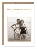 Two women in vintage swimsuits stand arm-in-arm in shallow ocean water, smiling. Above them, a quote reads, "Happy girls are the prettiest," attributed to Audrey Hepburn‚Äîpart of the award-winning Happy Girls Birthday Card collection by Shannon Martin Design that captures timeless joy.