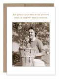 The Apple A Day Get Well Card by Shannon Martin Design features a vintage-style photo of a smiling person sitting next to a basket of apples and holding one, with the humorous caption above: "An apple a day will keep anyone away, if thrown hard enough." This card is printed on recycled paper and proudly made in the USA.