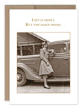 A vintage photo of a woman in a coat and hat standing next to an old car, with another person visible inside. The text above reads, "Life is short. Buy the damn shoes." It's reminiscent of flipping through sepia-colored memories, much like the nostalgic essence captured in the *Buy The Shoes Birthday Card* by *Shannon Martin Design*.