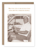 A charming black and white image shows a senior gentleman seated in a car, happily gripping the steering wheel. Adorned with a white cap and glasses, he exudes a warm smile. The card features the text: "May you live to be so old that your driving terrifies people." This delightful card is crafted on recycled paper by Shannon Martin Design and is part of their "Driving Terrifies" Birthday Card collection.