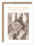 A birthday card from Shannon Martin Design's collection, "Into This World Birthday Card," features a black-and-white image of a woman in a party hat holding a cocktail glass. The card humorously advises celebrating "the way you came into this world, naked and screaming." It is crafted on recycled paper with vintage photos and balloons decorating the background.