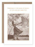 A sepia-toned image from the Shannon Martin Design winds encouragement card portrays a woman outdoors holding a windblown umbrella. She is dressed in a gingham dress, with the words above stating, "Sometimes in the winds of change, we find our true direction.