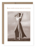 The "Being Happy Birthday Card" by Shannon Martin Design is a vintage card crafted from recycled paper and showcases a sepia-toned photo of a woman joyfully posing in a long skirt and blouse against a cloudy sky. The caption reads, "Being happy never goes out of style.