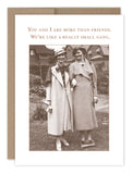 In a garden, two women dressed in vintage clothing smile while holding blank vintage photos. Above them is the text: "You and I are more than friends. We're like a really small gang." Their hats and coats add a nostalgic and humorous touch, perfectly captured by the Small Gang Friendship / Just Because Card from Shannon Martin Design.