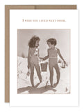 Two young girls in vintage swimsuits stand hand-in-hand on a sandy beach, each holding a small bucket. The card, titled "Next Door Friendship / Just Because Card" by Shannon Martin Design, is printed on FSC-certified recycled paper and bears the message, "I wish you lived next door." It fits snugly into Shannon's signature Kraft envelope, adding the perfect nostalgic touch.