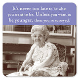 A black and white photo features an elderly woman sitting at a table with Shannon Martin Design's "Never Too Late" Coaster in front of her. She's wearing glasses and a lacy shawl. The text above reads: It's never too late to be what you want to be. Unless you want to be younger, then you're screwed.