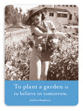 A black-and-white photo features a woman in a wide-brimmed hat standing in a garden, holding a basket of flowers. Below the image is a blue banner displaying Audrey Hepburn's inspiring quote, "To plant a garden is to believe in tomorrow." This design from Shannon Martin Design makes for the perfect addition to your collection of fridge magnets under the name "To Plant A Garden Magnet.