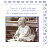An elderly woman sits on a chair, wearing glasses and a knitted shawl, holding a pen and paper. The text above reads, "It‚Äôs never too late to be what you want to be. Unless you want to be younger, then you‚Äôre screwed." Pair it with Shannon Martin Design's eco-friendly Never Too Late Beverage Napkin for an added touch!